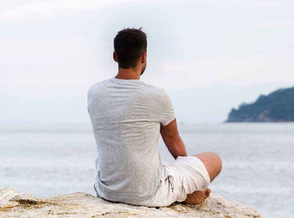 A man sits on a rock, gazing out at the vast ocean under a clear blue sky.
